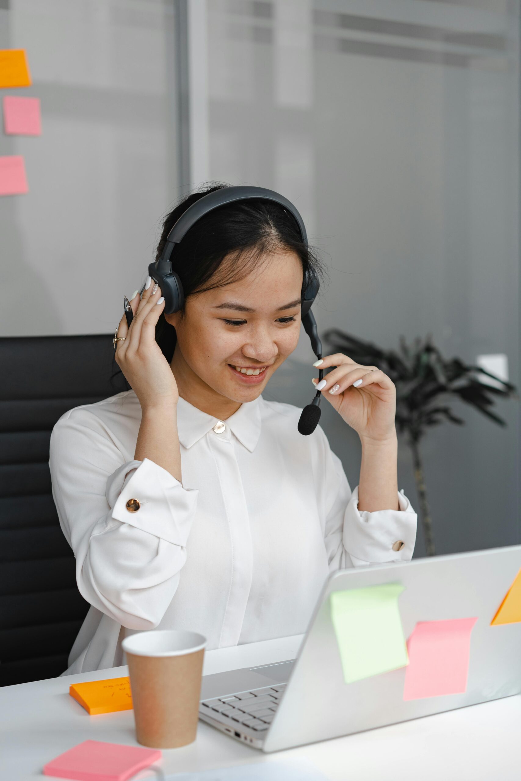 Smiling Asian woman in white shirt working as a call center agent with laptop and headset in office.